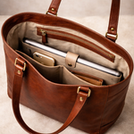 Brown leather handbag with compartments holding a laptop and phone on a beige background